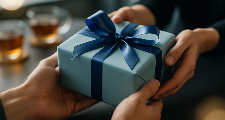 Nahaufnahme von Händen, die ein ordentlich verpacktes Geschenkpaket mit einer blauen Schleife austauschen, mit zwei Tassen Tee im unscharfen Hintergrund auf einem dunklen Tisch.