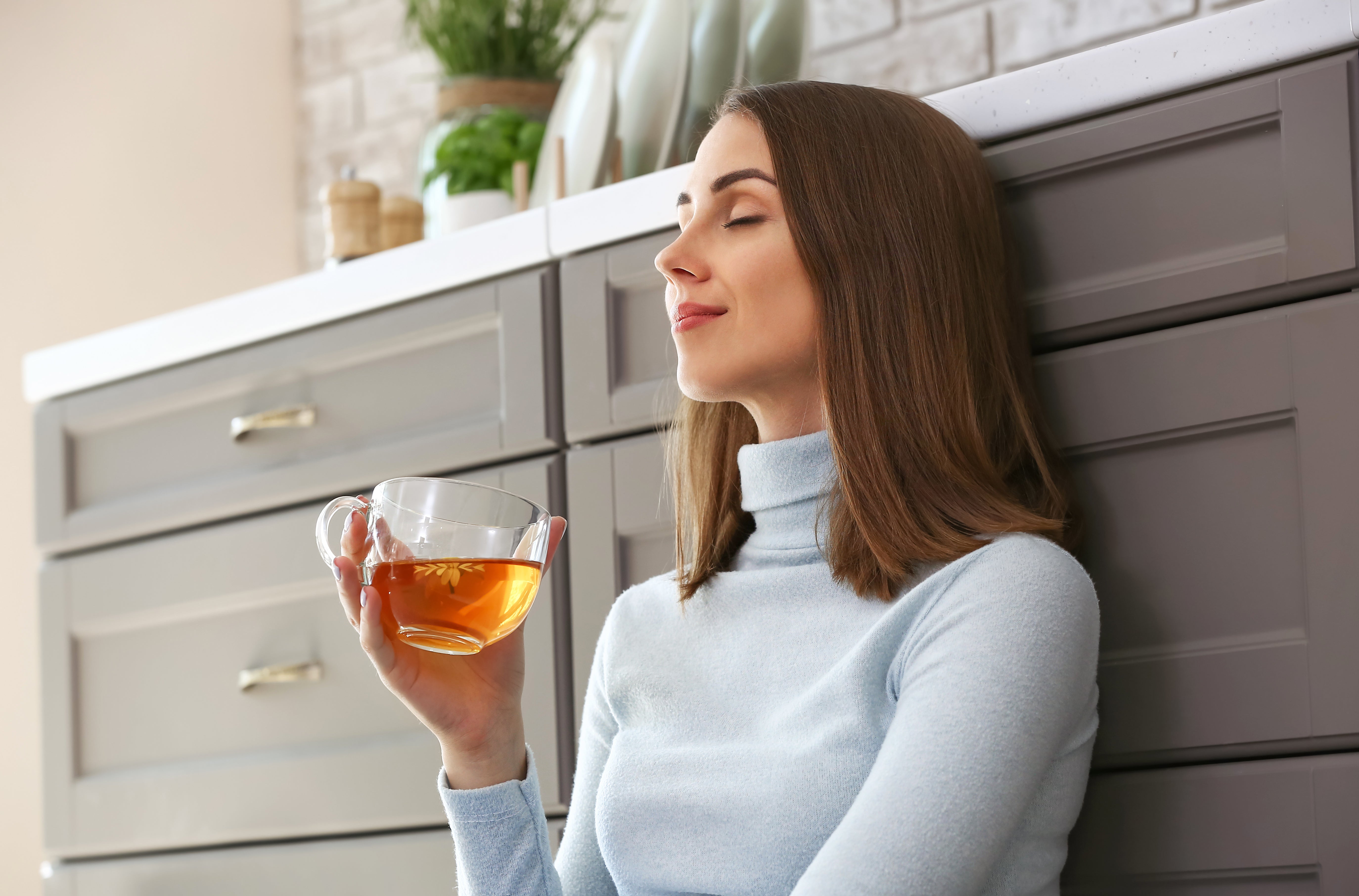 Eine Frau mit lockigem Haar lächelt mit geschlossenen Augen, während sie eine Glastasse mit rotem Tee in der Hand hält. Im Hintergrund ist ein geschmückter Weihnachtsbaum zu sehen.