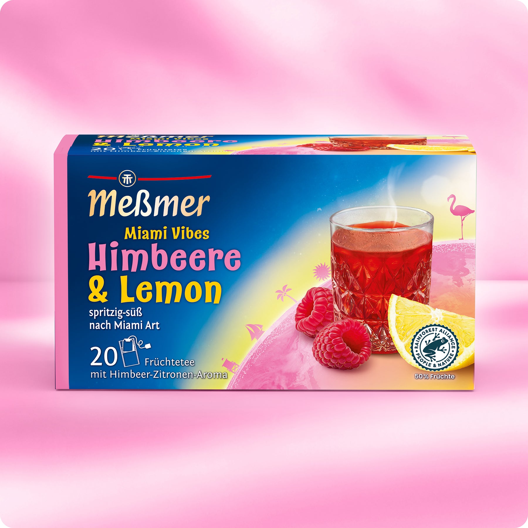 Eine farbenfrohe Schachtel Meßmer DE Miami Vibes Tee steht auf einer rosafarbenen Fläche und zeigt roten Tee in einem Glas, Himbeeren, Zitronenscheiben und Flamingos vor einem lebhaften tropischen Hintergrund.