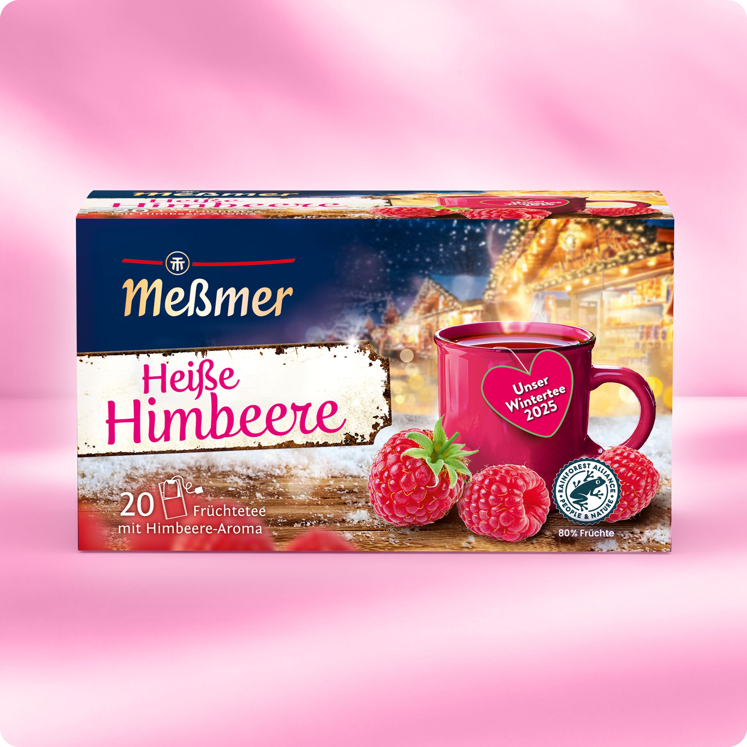 Eine Schachtel Meßmer DE Heiße Himbeere Früchtetee steht auf einer rosafarbenen Fläche, die Himbeeren, eine rote Teetasse und eine festliche Wintermarktszene zeigt. Die Verpackung enthält 20 Teebeutel.