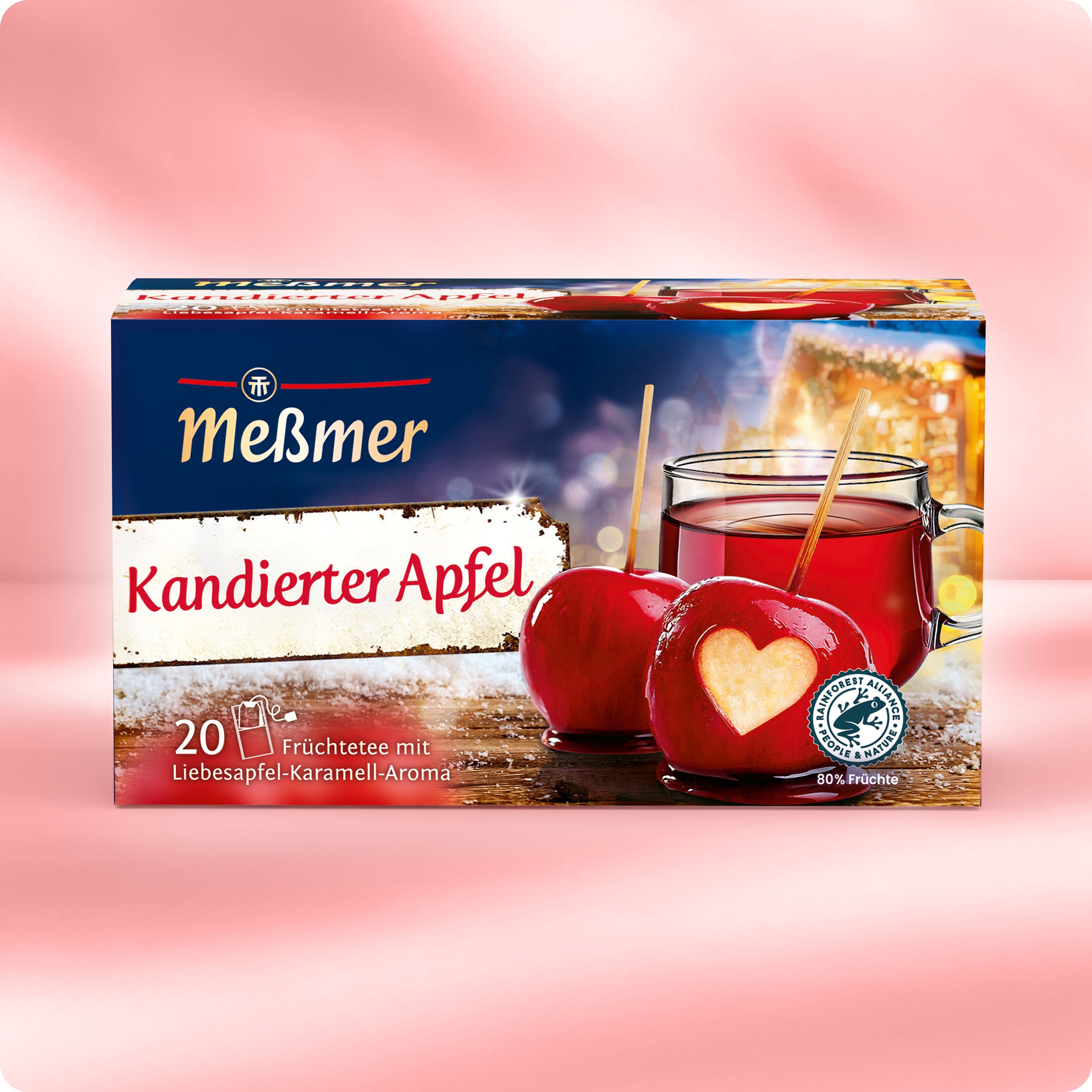 Eine Schachtel Meßmer DE Kandierter Apfel ist auf einem rosa Hintergrund abgebildet. Auf der Verpackung sind zwei rote kandierte Äpfel, eine Teetasse und herzförmige Apfelspalten abgebildet, die winterliches Weihnachtsmarkt-Ambiente in Ihr Zuhause bringen.