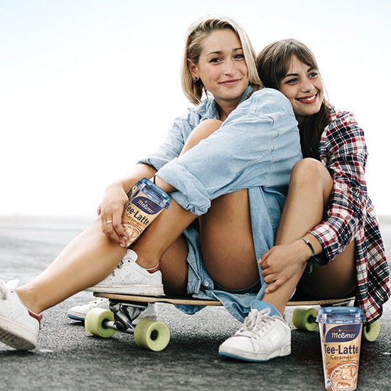 Zwei lächelnde junge Frauen sitzen im Freien auf einem Skateboard, die eine im Jeanshemd, die andere im karierten Hemd, und halten jeweils einen Meßmer Tee-Latte Karamelldrink in der Hand. Im Vordergrund ist auch ein Behälter zu sehen.