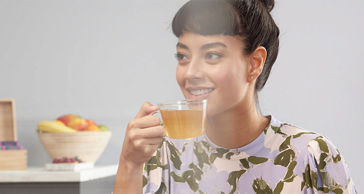 Eine Frau mit dunklem Haar in einem Dutt und einem geblümten Oberteil lächelt, während sie eine Glastasse mit Tee hält. Im Hintergrund stehen eine Schale mit Obst und eine Holzkiste auf einer hellen Arbeitsplatte.
