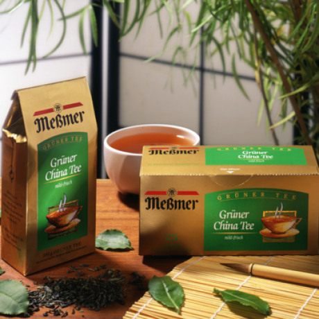 Kisten mit Meßmer Grüner China Tee werden mit losen Teeblättern, einer Bambusmatte, frischen grünen Blättern und einer Tasse Tee auf einem Tisch präsentiert, mit Pflanzen und einem Reispapierschirm im Hintergrund.