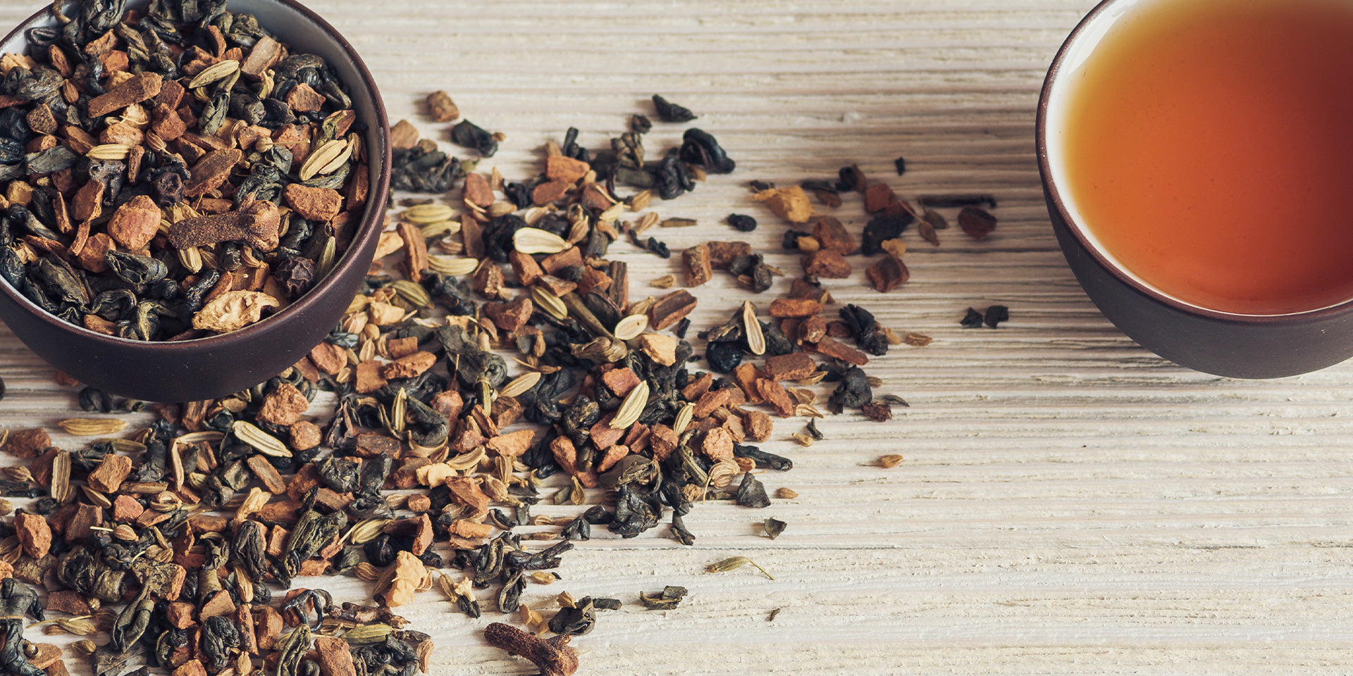 Eine Schale mit losem Blatttee und Gewürzen steht auf einer hellen Holzfläche neben einer Tasse mit aufgebrühtem Tee von warmer Bernsteinfarbe.