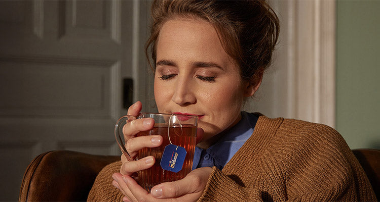 Eine Frau mit geschlossenen Augen lächelt sanft, während sie mit beiden Händen eine durchsichtige Tasse Tee hält. Sie trägt einen braunen Pullover und ein blaues Hemd und sitzt in einem Haus neben einer grauen Tür.