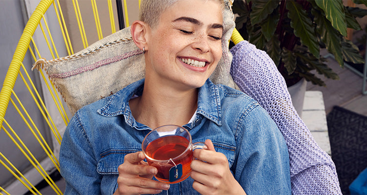 Eine Person mit kurzen blonden Haaren, die ein Jeanshemd trägt, lächelt mit geschlossenen Augen und hält eine Tasse Tee in der Hand. Sie sitzt bequem auf einem gelben Stuhl mit einem Kissen und einer lila Strickdecke.
