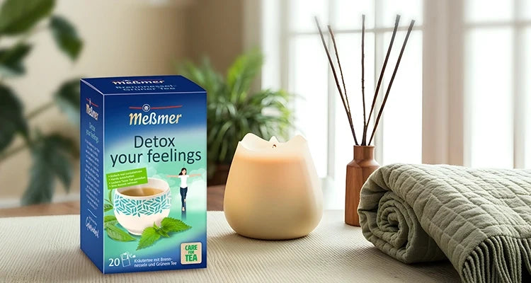 Eine Schachtel Meßmer Gelassenheit-Tee steht auf einer hellvioletten Fläche mit Kerzen, einer Tasse Tee, Dekosteinen und einer Strickdecke im Hintergrund.