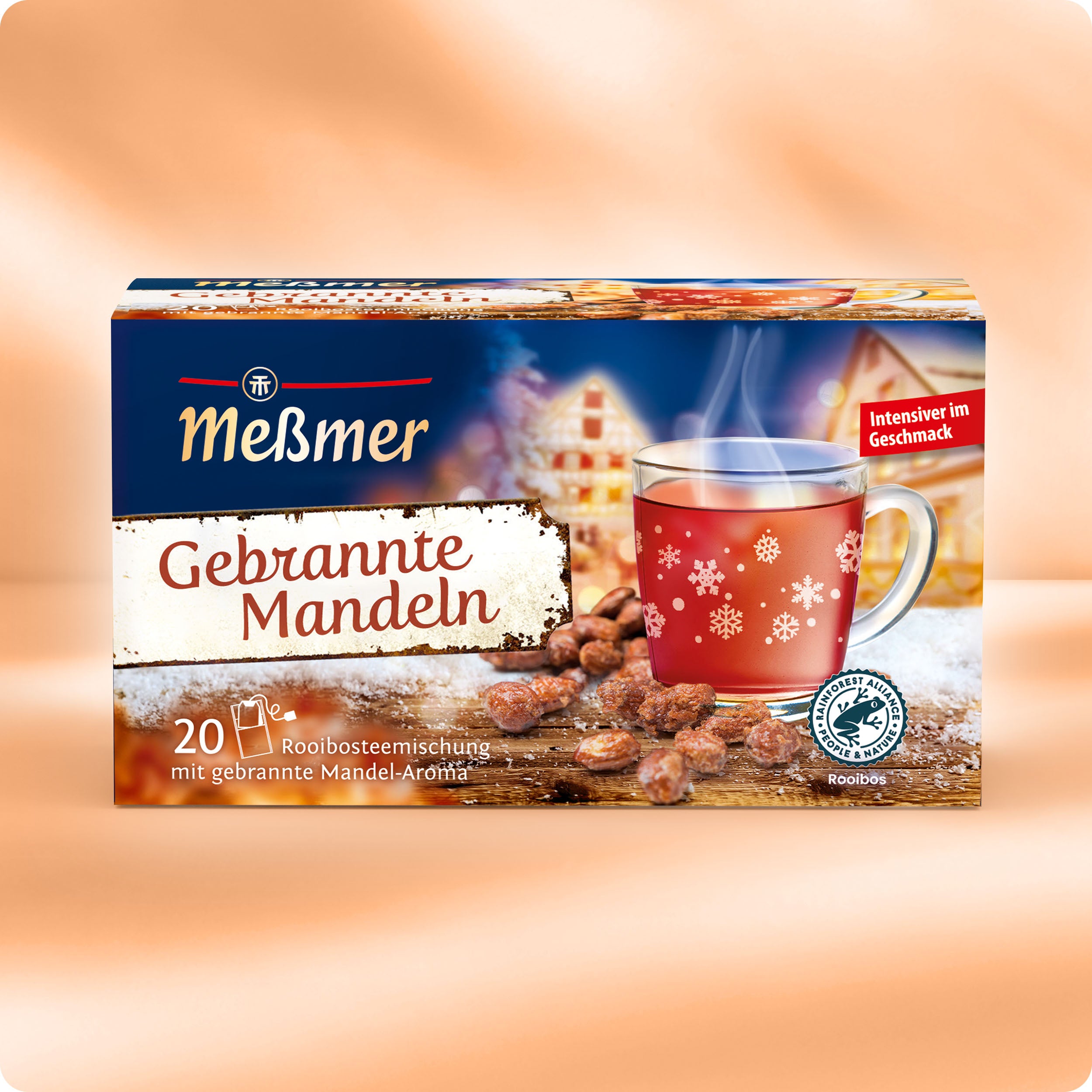 Eine Schachtel Meßmer DE Gebrannte Mandel Tee wird mit einer Glastasse mit rotem Tee, Schneeflockenmotiven, gerösteten Mandeln und einem festlichen Winterhintergrund gezeigt. Die Schachtel enthält 20 Rooibos-Teebeutel mit Mandelaroma.
