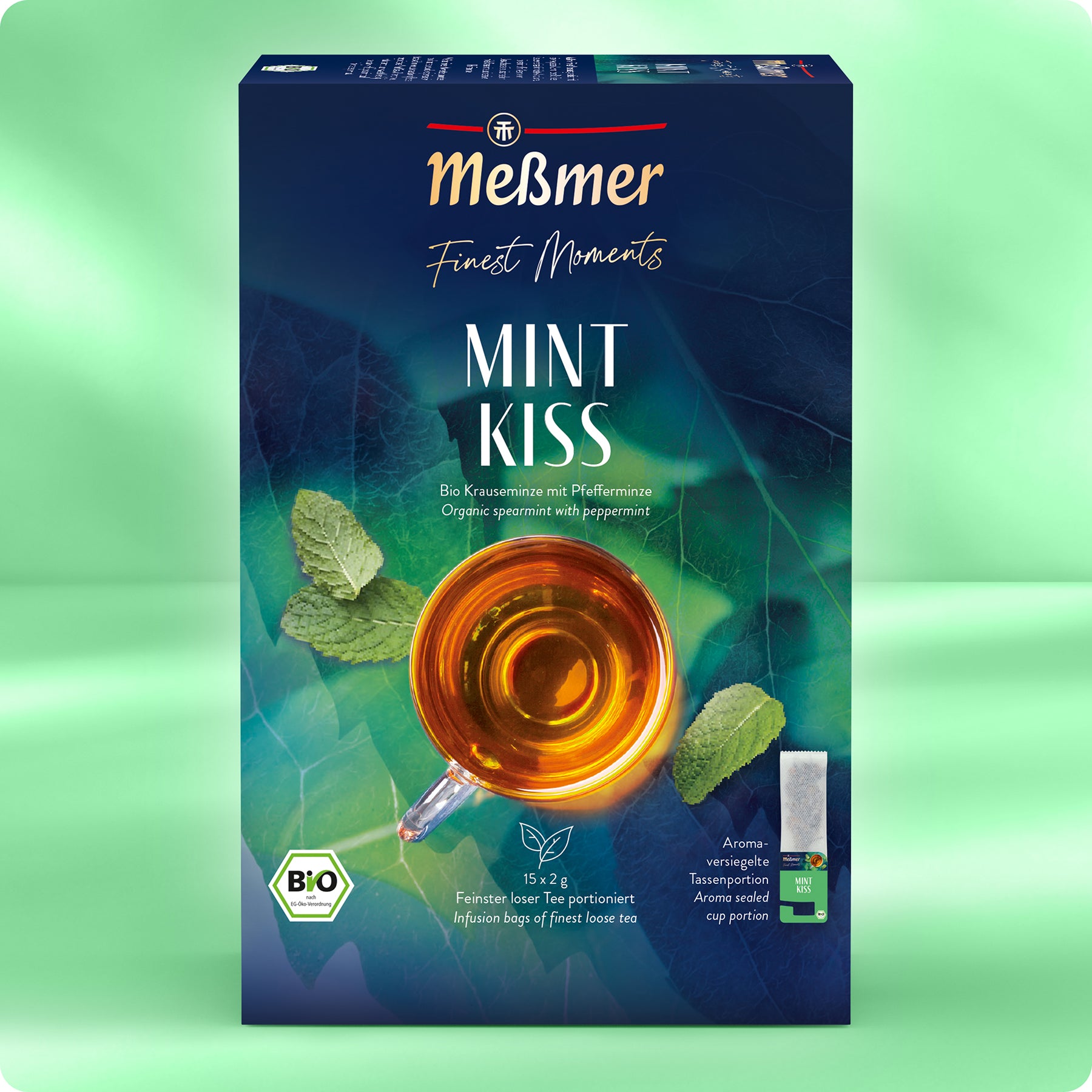 Eine Schachtel Meßmer DE Bio Mint Kiss besteht aus einer dunkelgrünen und blauen Verpackung mit dem Produktnamen in weißer Farbe, einer Glastasse mit Tee, Minzblättern, einem BIO-Etikett und einer Abbildung von Kräuter-Früchtetee.