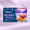 Verpackung von Wintermagie mit Früchtetee in Tasse, Spekulatiusgebäck, Orangenscheiben vor winterlicher Waldlandschaft und Care-for-Tea-Siegel für nachhaltige Zutaten.