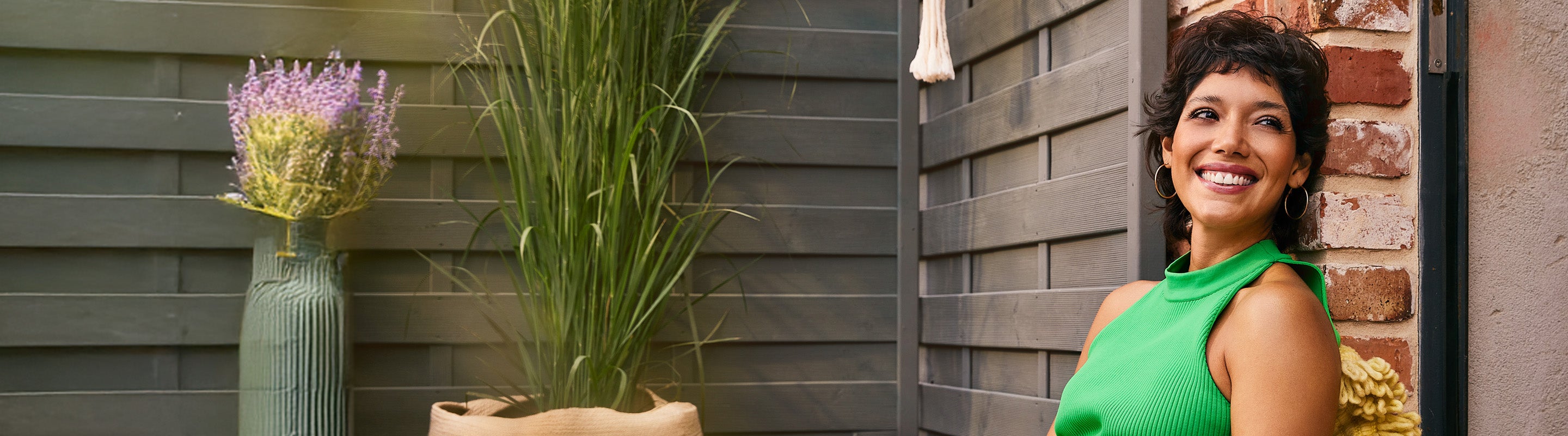 Eine Frau in einem hellgrünen, ärmellosen Oberteil sitzt lächelnd an einer Backsteinmauer, neben ihr stehen hohe grüne Pflanzen und lila Blumen in Vasen, in einem Außenbereich.