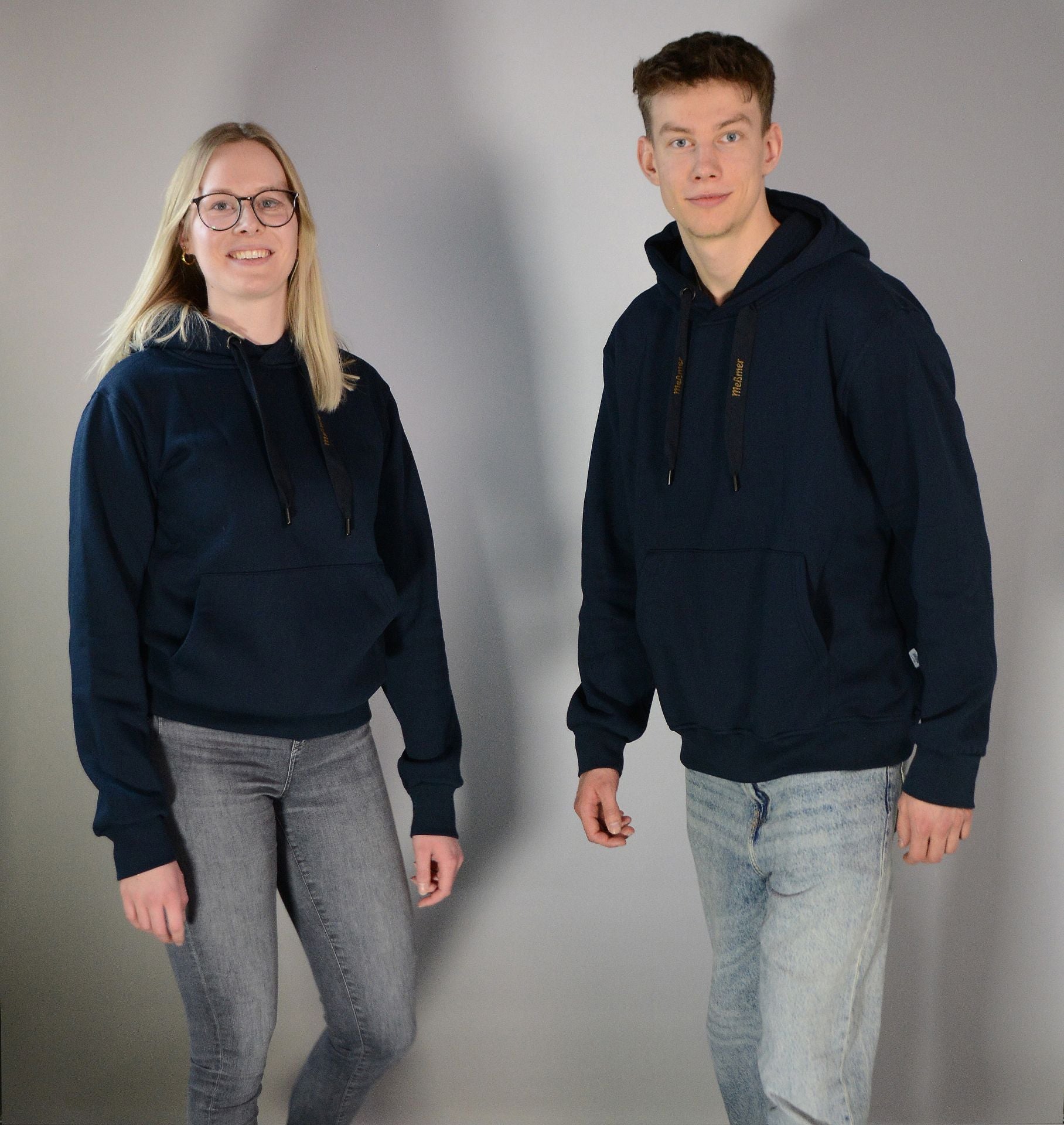 Zwei Personen in gemütlichen Meßmer DE Hoodie Unisex Kapuzenpullis und hellen Jeans posieren vor einem schlichten, hellgrauen Hintergrund. Die eine links hat lange blonde Haare und eine Brille, die andere hat kurze Haare. Beide lächeln.