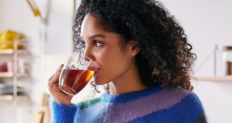 Eine Frau mit lockigem Haar und einem blau-lila Pullover trinkt Tee aus einem klaren Glasbecher. Sie blickt zur Seite in einer hell erleuchteten, modernen Küche.